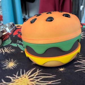 May include: A colorful, toy hamburger with an orange top bun, green lettuce, a dark brown patty, and a yellow bottom bun. The top bun has black sesame seeds. The burger is sitting on a black patterned surface.