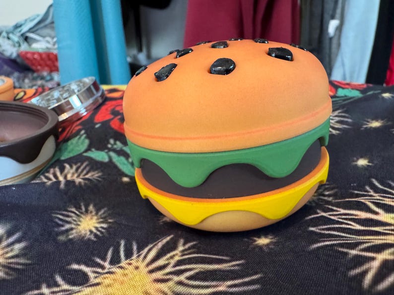 May include: A colorful, toy hamburger with an orange top bun, green lettuce, a dark brown patty, and a yellow bottom bun. The top bun has black sesame seeds. The burger is sitting on a black patterned surface.