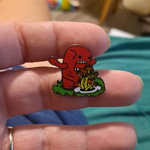 May include: Red cartoon monster with a wide open mouth eating a bunch of brown mushrooms. The monster is standing on green grass and there is a white plate under the mushrooms.