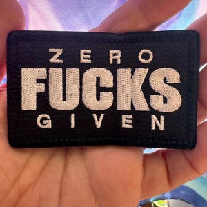 May include: A black rectangular patch with white embroidered text that reads "ZERO FUCKS GIVEN". The patch is held in a hand against a colorful, blurred background.