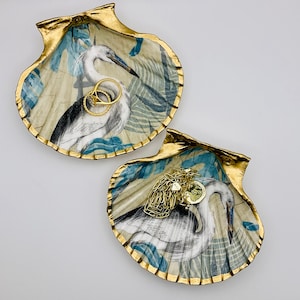 May include: Two decorative seashell-shaped trinket dishes with gold trim. Each dish features a white egret bird with blue and green tropical leaf patterns. One dish has two gold rings inside, and the other has a gold chain necklace.