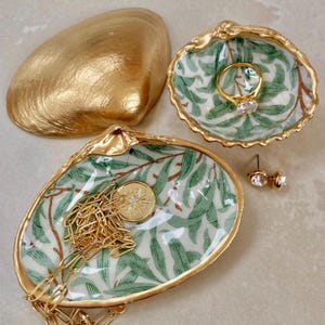 May include: A collection of decorative seashell jewelry dishes. One large gold-painted shell and two smaller shell dishes with a green and white leaf pattern and gold trim. A gold ring, earrings, and necklace are displayed.