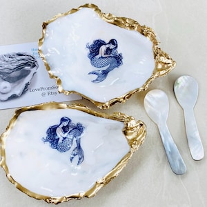 May include: Two white oyster shell dishes with gold rims and blue mermaid illustrations. Two mother-of-pearl spoons are next to the dishes. The text "LoveFromSo @ Etsy" is visible in the background.