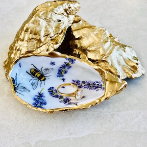 May include: A gold-painted oyster shell ring dish. The interior features a white background with bee and lavender illustrations. A gold ring with a large, clear gemstone sits inside the dish.
