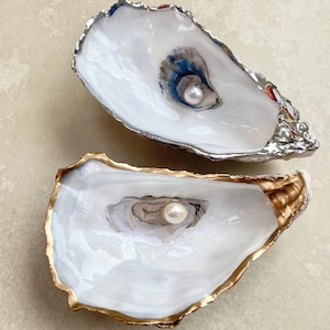 May include: Two decorative oyster shell dishes. One has a silver rim and a blue and grey interior, the other has a gold rim. Each shell contains a pearl in the centre.