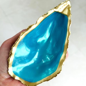 May include: A decorative oyster shell dish featuring a vibrant, glossy turquoise interior and a metallic gold painted rim. The shell has an irregular, organic shape with a smooth, reflective surface and a textured, gilded edge that follows the natural contour.