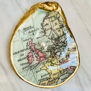 May include: An oyster shell with a gold trim, decorated with a vintage map of Europe and a pearl. The shell is about 10 cm long and 7.6 cm wide, suitable for holding small trinkets or jewellery.