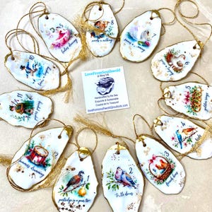 May include: Twelve oyster shell ornaments featuring the Twelve Days of Christmas illustrations, each with gold-painted edges and gold hanging strings. A central square card displays the text LoveFromSouthwold at Etsy UK and descriptive business details.