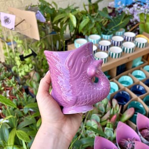 May include: A lilac ceramic planter shaped like a mythical creature, possibly a dragon or fish, with detailed scales and fins. The planter is held in a hand, with other small ceramic pots in the background.