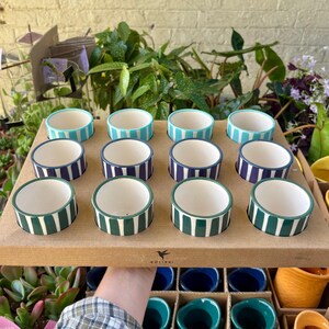 May include: A wooden tray displays twelve small ceramic bowls. Each bowl has a white interior and a striped exterior in shades of teal, navy, and green. The bowls are arranged in three rows of four.