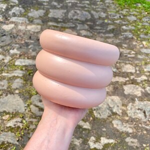 May include: A pink ceramic planter with a unique, wavy, layered design. The planter is made of smooth, textured ceramic and has a light pink colour.