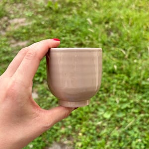 May include: A small, beige ceramic cup with a rounded body and a slightly flared base. The cup is held against a backdrop of green grass. The cup's smooth surface and neutral colour suggest a minimalist design, suitable for beverages.