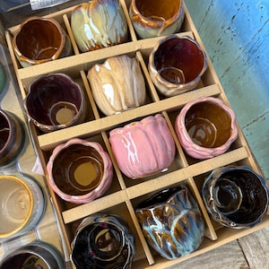 May include: A collection of ten small ceramic cups with scalloped edges, arranged in a cardboard grid. The cups feature various glossy glaze finishes in shades of pink, brown, tan, blue, and black, with some showing multi-coloured marbling.