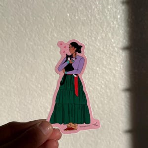 May include: A sticker featuring an illustration of a person holding a black and white cat. The person is wearing a purple top, green skirt, and red sash. Pink hearts float above the person's head.