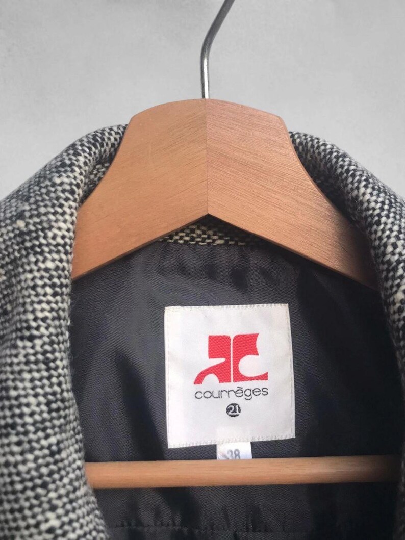 May include: A black and white tweed jacket hanging on a wooden hanger. The jacket has a black lining and a label with the Courr&egrave;ges logo in red and the number 21. The size tag reads 38.