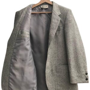 May include: A light gray tweed blazer with a notched lapel, two front pockets, and two black buttons on the sleeve. The blazer is open, revealing a gray satin lining and a small interior pocket. It hangs on a wooden hanger.