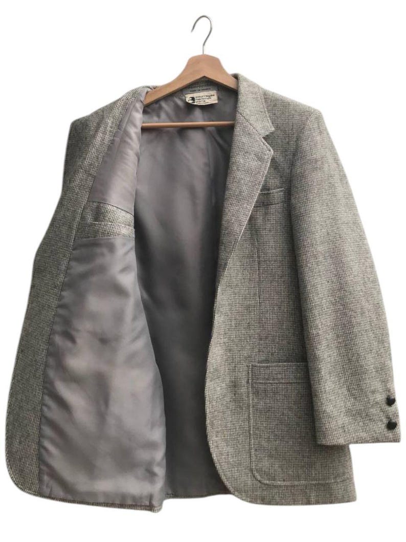 May include: A light gray tweed blazer with a notched lapel, two front pockets, and two black buttons on the sleeve. The blazer is open, revealing a gray satin lining and a small interior pocket. It hangs on a wooden hanger.
