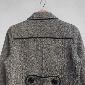 May include: A gray tweed coat with black trim and buttons. The coat has a classic design with a notched collar and a decorative flap detail on the back. The fabric has a speckled pattern, and the coat is hanging on a wooden hanger.