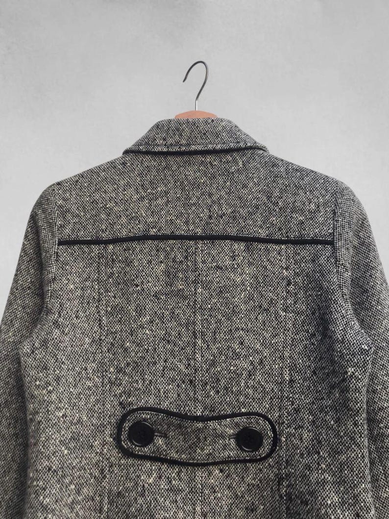 May include: A gray tweed coat with black trim and buttons. The coat has a classic design with a notched collar and a decorative flap detail on the back. The fabric has a speckled pattern, and the coat is hanging on a wooden hanger.