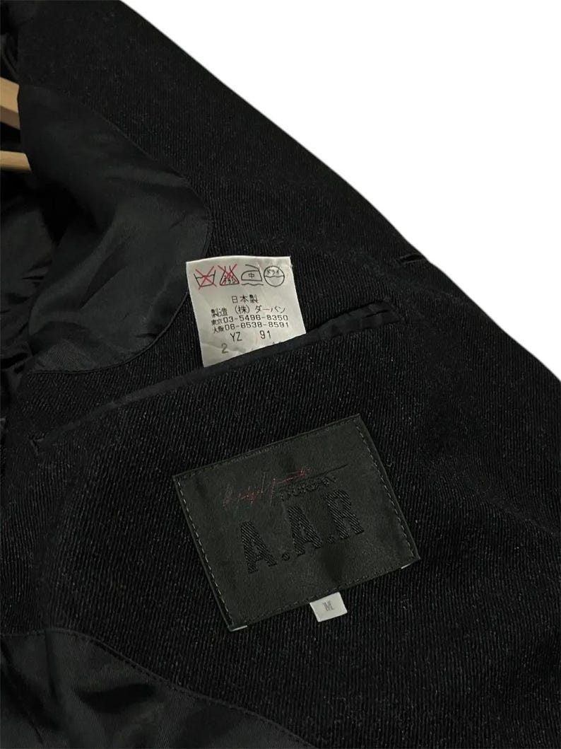 K&ouml;nnte beinhalten: Nahaufnahme des Innenraums einer schwarzen Wolljacke. Das Futter ist schwarz, und ein Etikett mit dem Text "A.A.R" ist sichtbar. Ein kleineres Etikett mit japanischem Text und Pflegesymbolen ist ebenfalls vorhanden. Die Jacke scheint Gr&ouml;&szlig;e M zu haben.