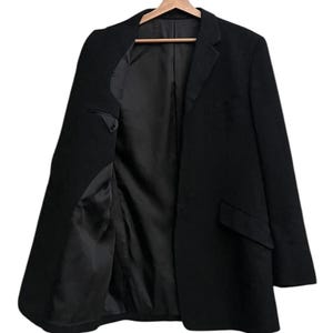 K&ouml;nnte beinhalten: Ein schwarzer Blazer h&auml;ngt an einem Holzb&uuml;gel. Der Blazer ist offen und zeigt das schwarze Futter und die Innentaschen. Die Jacke hat ein klassisches Revers und zwei Vordertaschen.