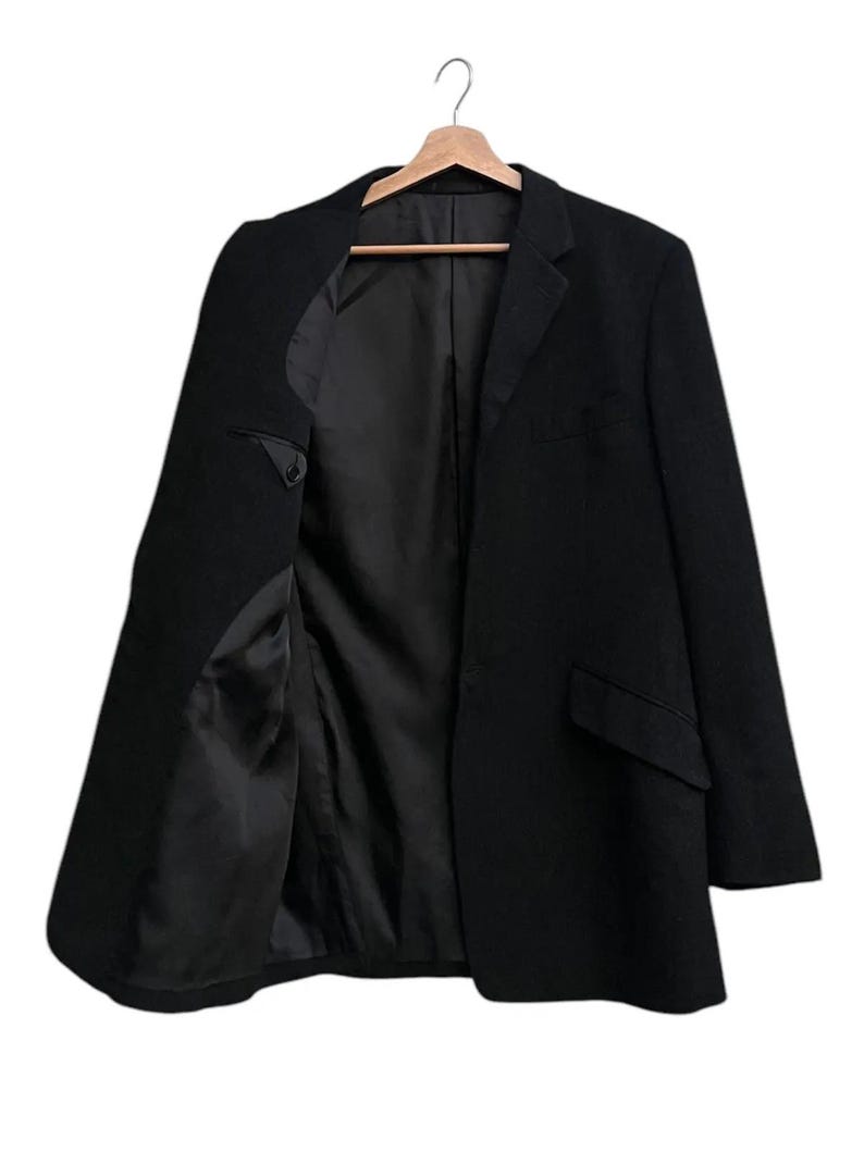 K&ouml;nnte beinhalten: Ein schwarzer Blazer h&auml;ngt an einem Holzb&uuml;gel. Der Blazer ist offen und zeigt das schwarze Futter und die Innentaschen. Die Jacke hat ein klassisches Revers und zwei Vordertaschen.
