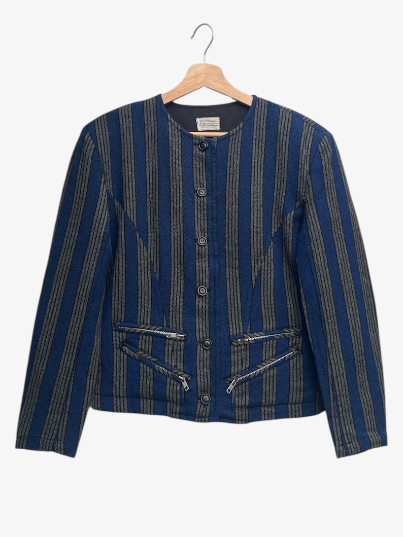 May include: A blue and gray striped jacket with a round collar and four buttons down the front. The jacket has two zippered pockets and is hanging on a wooden hanger.