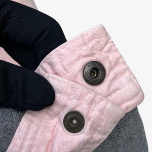 Puede incluir: Primer plano de una prenda gris con cuello rosa claro. El cuello se asegura con dos cierres de bot&oacute;n met&aacute;licos. Los botones a presi&oacute;n tienen el texto "MICHE" impreso.