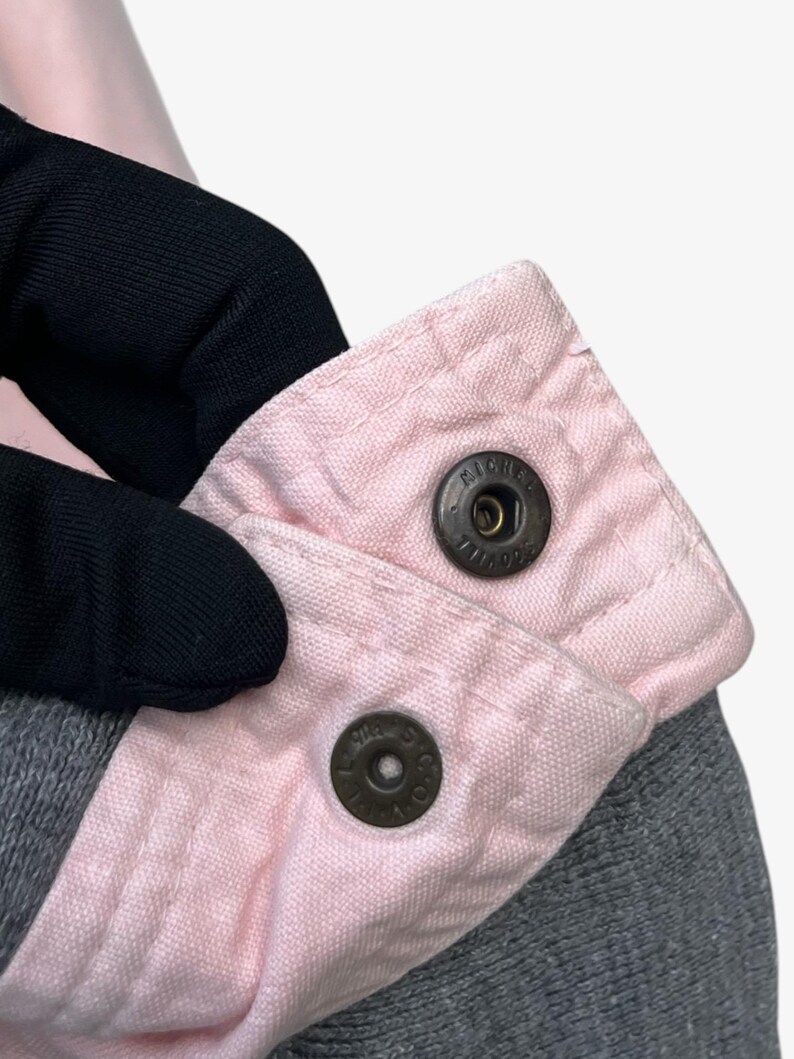 Puede incluir: Primer plano de una prenda gris con cuello rosa claro. El cuello se asegura con dos cierres de bot&oacute;n met&aacute;licos. Los botones a presi&oacute;n tienen el texto "MICHE" impreso.