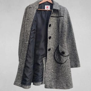 May include: A gray and white tweed coat with a black lining and black buttons. The coat has a classic design with a notched collar and a unique pocket detail. The coat is hanging on a wooden hanger.