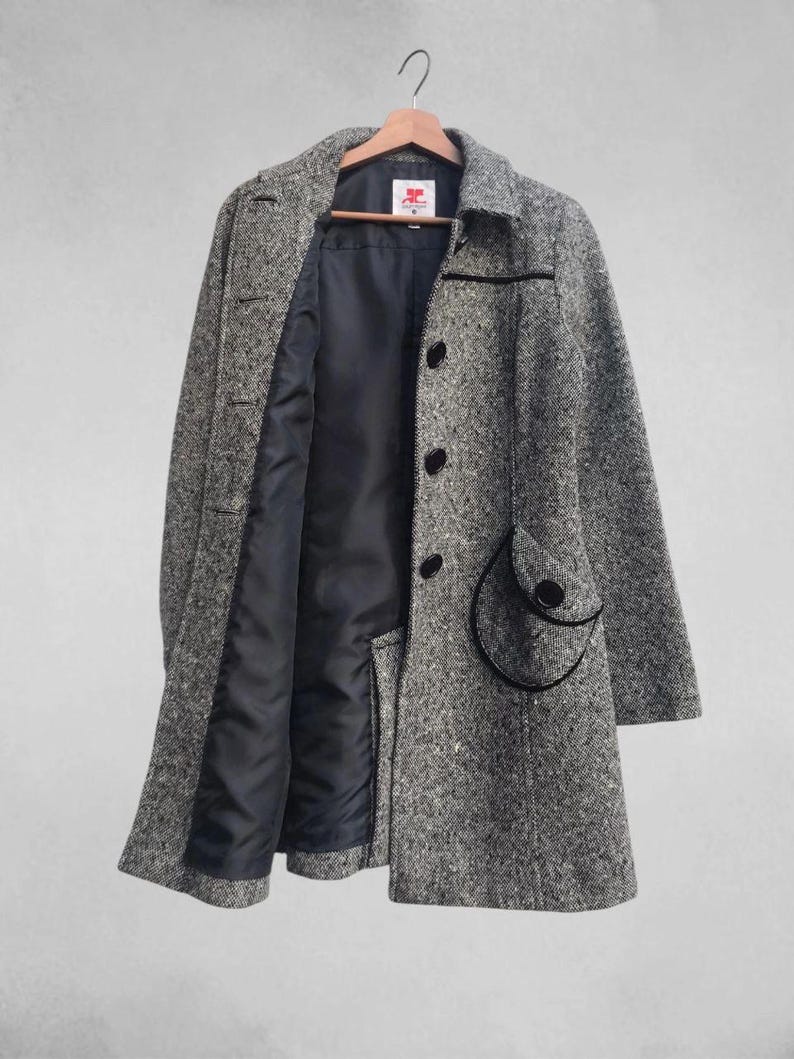 May include: A gray and white tweed coat with a black lining and black buttons. The coat has a classic design with a notched collar and a unique pocket detail. The coat is hanging on a wooden hanger.