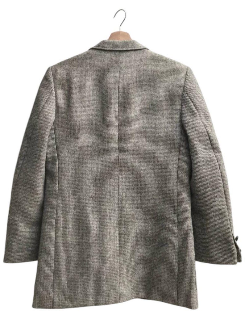 May include: A tweed blazer in a neutral color palette hangs on a wooden hanger. The jacket features a classic notched lapel, two buttons on the sleeves, and a tailored fit. The fabric has a subtle textured pattern.