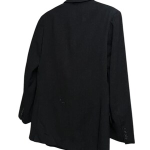 K&ouml;nnte beinhalten: Ein schwarzer Blazer h&auml;ngt an einem Holzkleiderb&uuml;gel. Der Blazer hat ein klassisches Design mit fallendem Revers und drei Kn&ouml;pfen an jeder Manschette. Die R&uuml;ckseite des Blazers zeigt die taillierte Passform und die Textur des Stoffes. Vielseitiges Kleidungsst&uuml;ck.