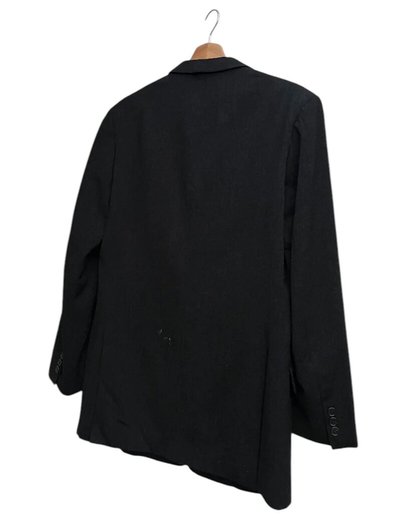 K&ouml;nnte beinhalten: Ein schwarzer Blazer h&auml;ngt an einem Holzkleiderb&uuml;gel. Der Blazer hat ein klassisches Design mit fallendem Revers und drei Kn&ouml;pfen an jeder Manschette. Die R&uuml;ckseite des Blazers zeigt die taillierte Passform und die Textur des Stoffes. Vielseitiges Kleidungsst&uuml;ck.