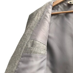 May include: Close-up of a light gray blazer with a textured, woven exterior. The interior lining is a smooth, solid gray. The blazer is hanging on a wooden hanger.