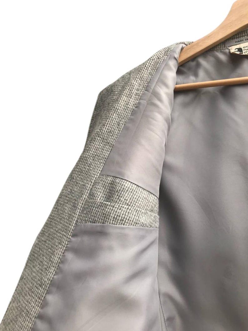 May include: Close-up of a light gray blazer with a textured, woven exterior. The interior lining is a smooth, solid gray. The blazer is hanging on a wooden hanger.