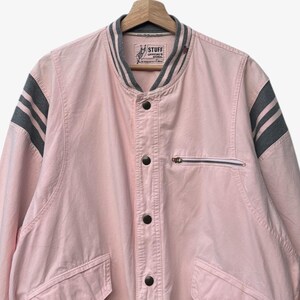 Puede incluir: Chaqueta bomber rosa claro con rayas grises en las mangas y el cuello. La chaqueta tiene un bolsillo con cremallera en el pecho y dos bolsillos delanteros. El texto "STUFF OFFICIAL'S SIGNAL" es visible en el cuello.