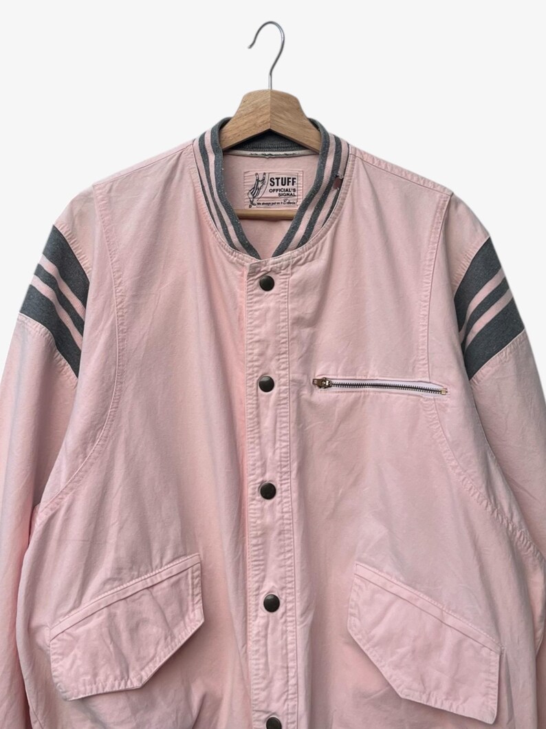 Puede incluir: Chaqueta bomber rosa claro con rayas grises en las mangas y el cuello. La chaqueta tiene un bolsillo con cremallera en el pecho y dos bolsillos delanteros. El texto "STUFF OFFICIAL'S SIGNAL" es visible en el cuello.