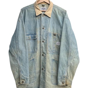 May include: Light blue denim jacket with a tan corduroy collar. The jacket features multiple pockets, button closures, and a faded, worn look. It is suspended from a wooden hanger.