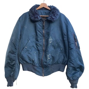 May include: A vintage blue bomber jacket with a faux fur collar. The jacket has a zip closure, ribbed cuffs, and a ribbed hem. There are some stains on the jacket. The jacket is hanging on a wooden hanger.
