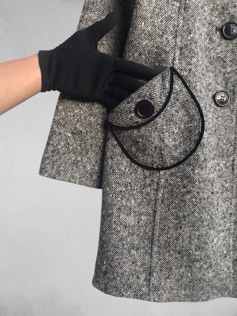 May include: A black and white tweed coat with a rounded pocket and black trim. The coat has large black buttons and a classic design. The sleeve is short and the fabric has a speckled pattern.