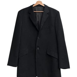 K&ouml;nnte beinhalten: Ein schwarzer Blazer mit zwei Vordertaschen und einem Zwei-Knopf-Verschluss, der an einem Holzb&uuml;gel h&auml;ngt. Der Blazer hat ein fallendes Revers und ein klassisches, ma&szlig;geschneidertes Design, das f&uuml;r verschiedene Anl&auml;sse geeignet ist.