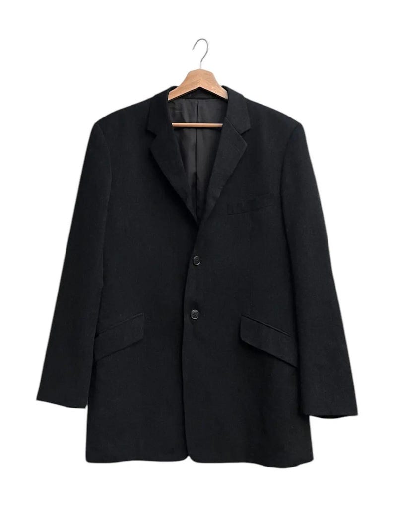 K&ouml;nnte beinhalten: Ein schwarzer Blazer mit zwei Vordertaschen und einem Zwei-Knopf-Verschluss, der an einem Holzb&uuml;gel h&auml;ngt. Der Blazer hat ein fallendes Revers und ein klassisches, ma&szlig;geschneidertes Design, das f&uuml;r verschiedene Anl&auml;sse geeignet ist.