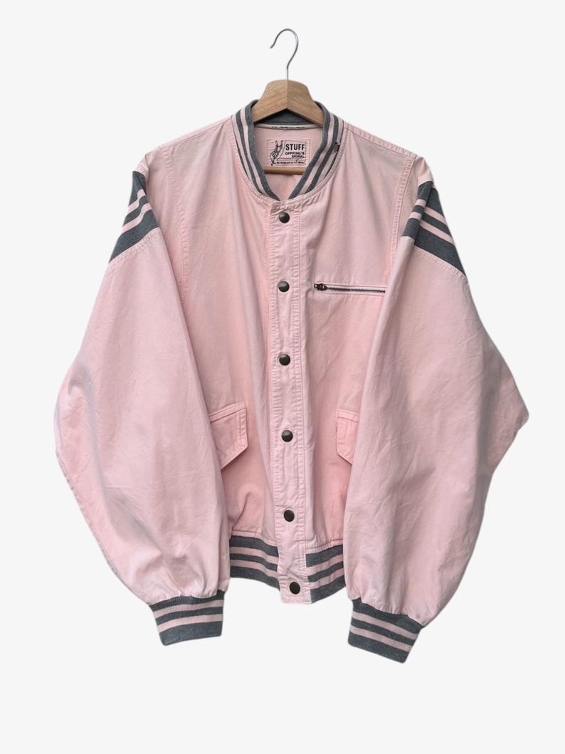Puede incluir: Chaqueta bomber rosa claro con rayas grises en las mangas y pu&ntilde;os. La chaqueta tiene cierre de botones, un peque&ntilde;o bolsillo con cremallera y la palabra "STUFF" en la etiqueta.