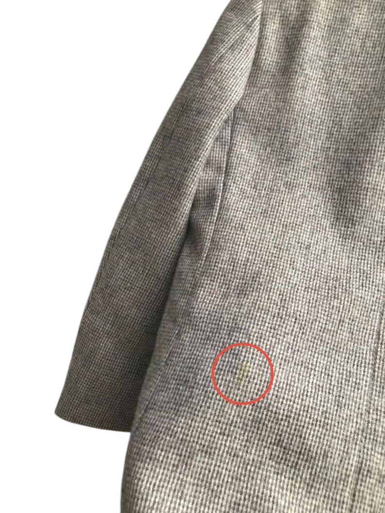 May include: Close-up of a gray blazer with a subtle houndstooth pattern. The fabric appears to be a woven material, with a visible sleeve and shoulder. A small stain is visible on the blazer.