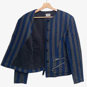 May include: A blue and gray striped jacket with a black interior, hanging on a wooden hanger. The jacket has a round collar, button closure, and two zippered pockets. The jacket's design features vertical stripes.