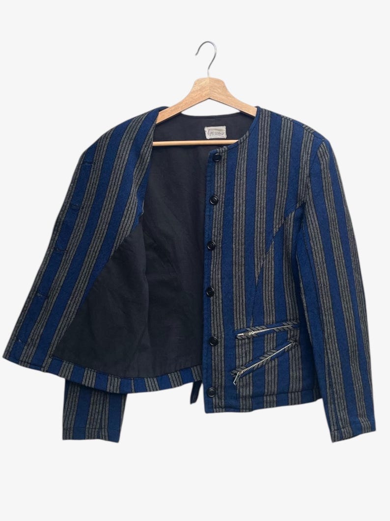 May include: A blue and gray striped jacket with a black interior, hanging on a wooden hanger. The jacket has a round collar, button closure, and two zippered pockets. The jacket's design features vertical stripes.