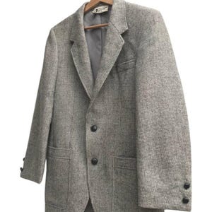 May include: A gray tweed blazer with a classic two-button closure, notched lapels, and two front pockets. The blazer is hanging on a wooden hanger. The sleeves have three buttons at the cuffs.