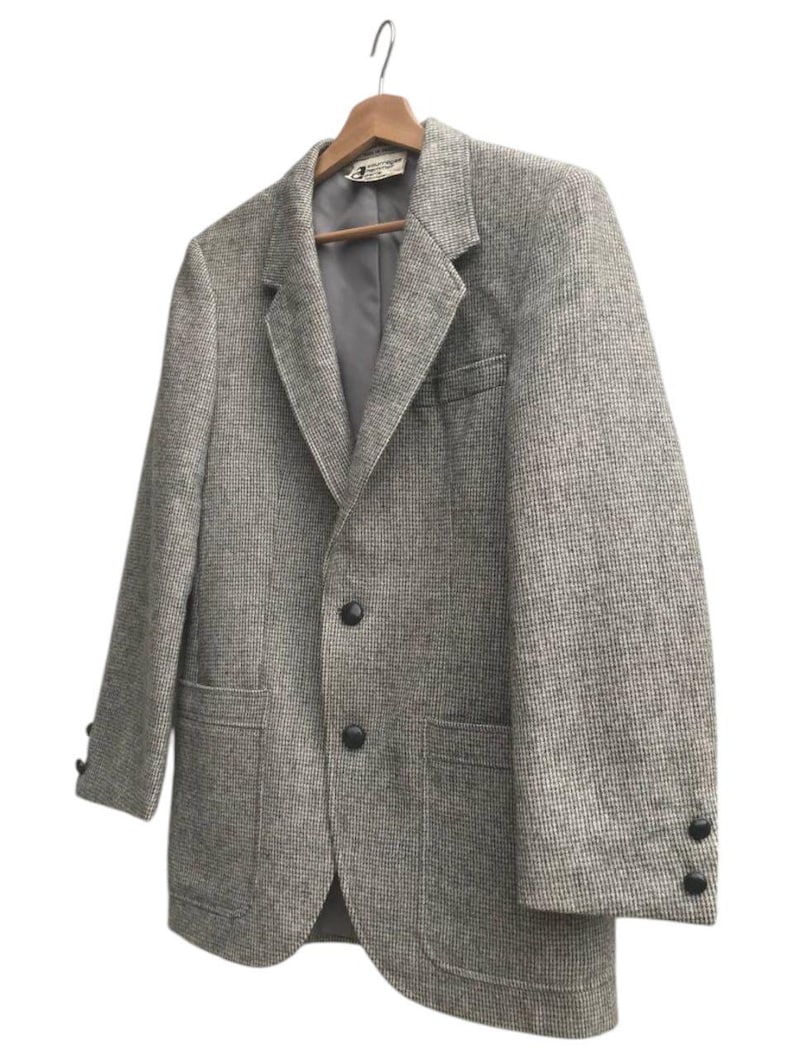 May include: A gray tweed blazer with a classic two-button closure, notched lapels, and two front pockets. The blazer is hanging on a wooden hanger. The sleeves have three buttons at the cuffs.