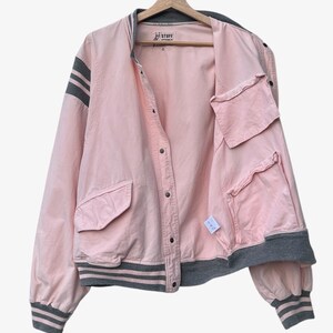 Puede incluir: Chaqueta bomber rosa con detalles grises, incluyendo rayas en las mangas y el ribete. La chaqueta tiene cierre de botones, bolsillos y la palabra "STUFF" en la etiqueta.